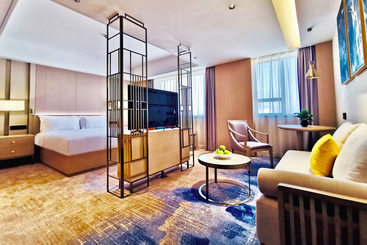 A modern hotel room with a bed, a TV, a seating area with a sofa and armchairs, and a small table with a plant.