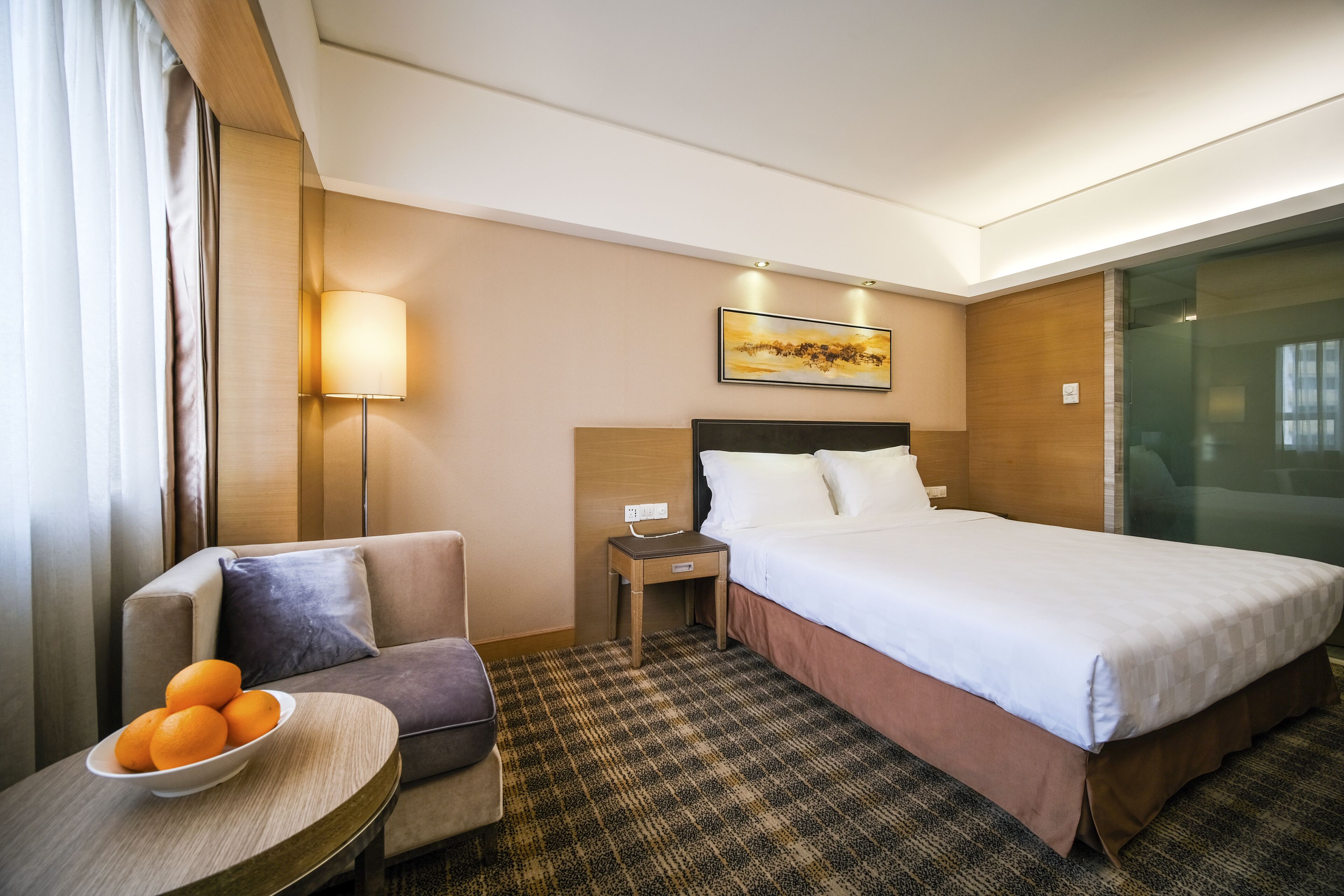 A hotel room with a bed, a nightstand, a lamp, a sofa, and a table with oranges.