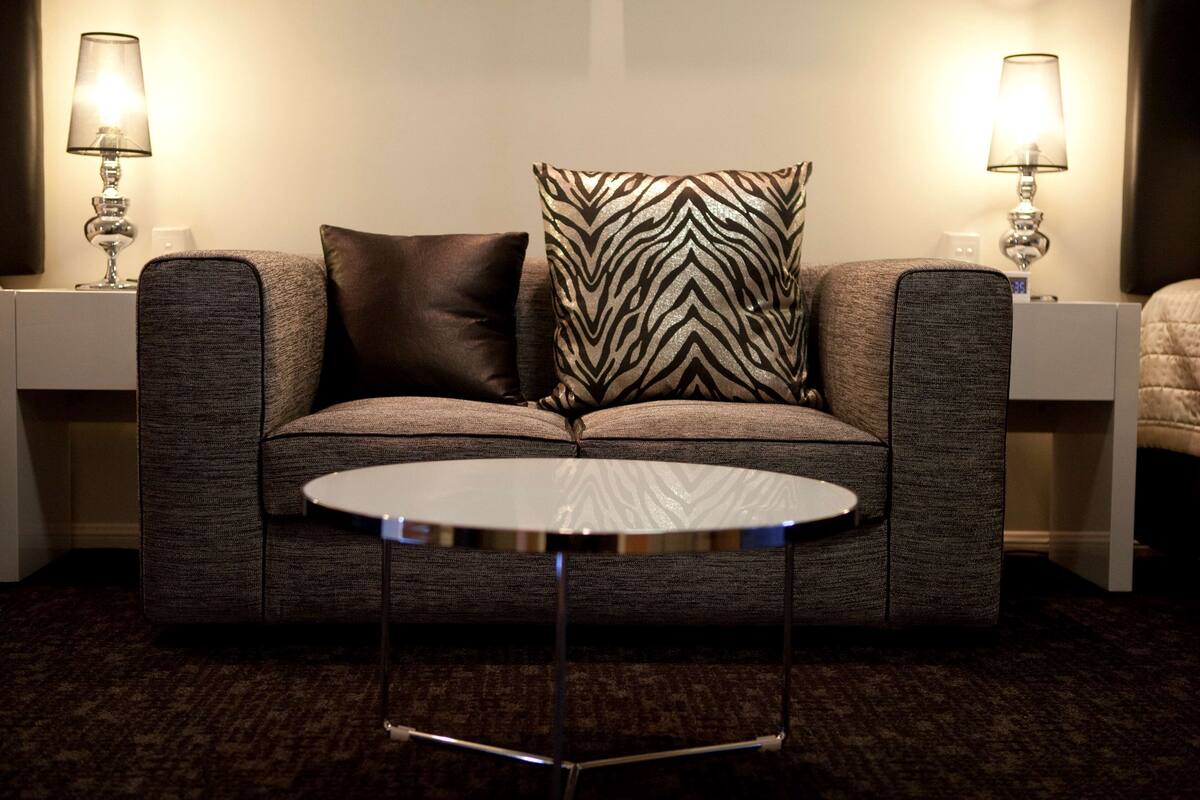 A modern hotel room with a grey sofa, two patterned pillows, a small round table, and two bedside lamps.