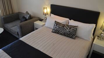 A hotel room with a bed, a sofa, a nightstand with a lamp, and a wall-mounted painting.
