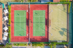 An aerial view of a tennis court with two courts, surrounded by a green fence and a covered seating area with umbrellas.