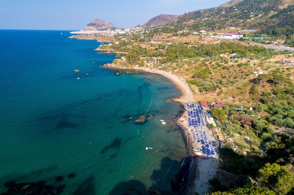 An aerial view of a coastal area with a beach, clear blue waters, and a line of blue umbrellas indicating a beach area.