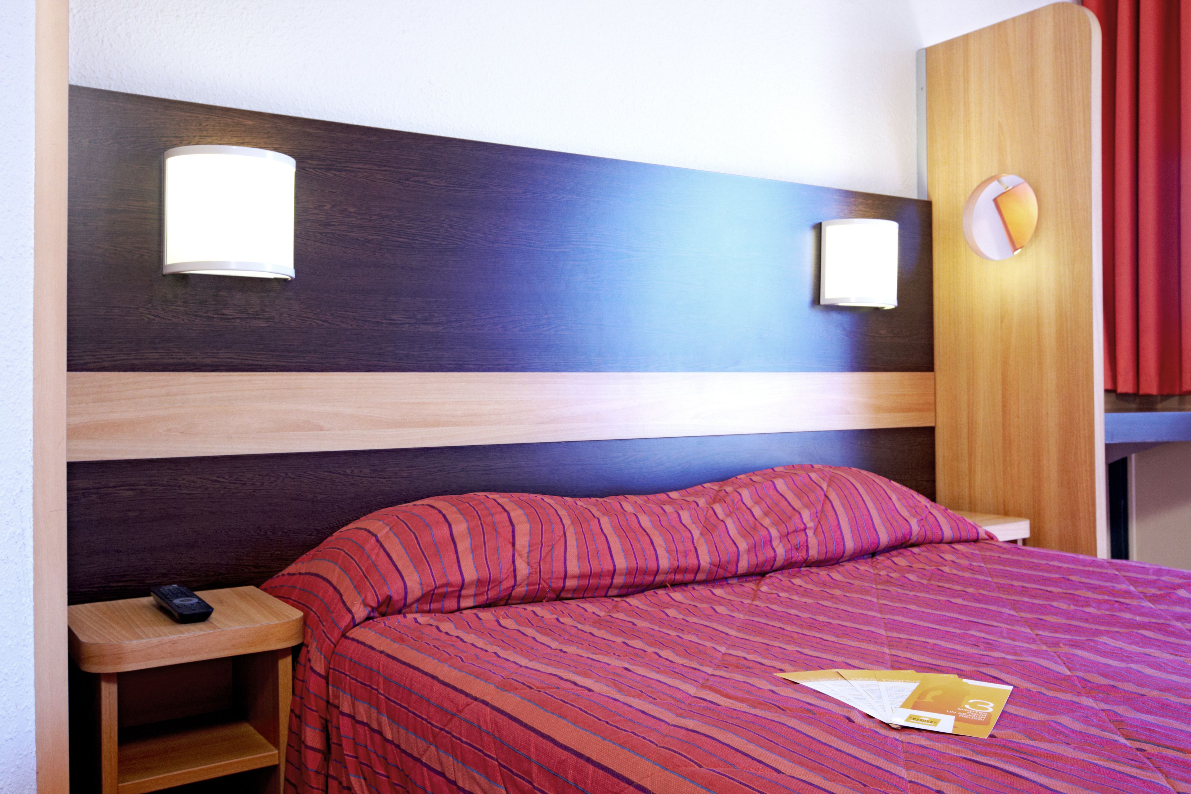 Quarto de hotel com cama, mesa de cabeceira, controle remoto e luminária fixa na parede.