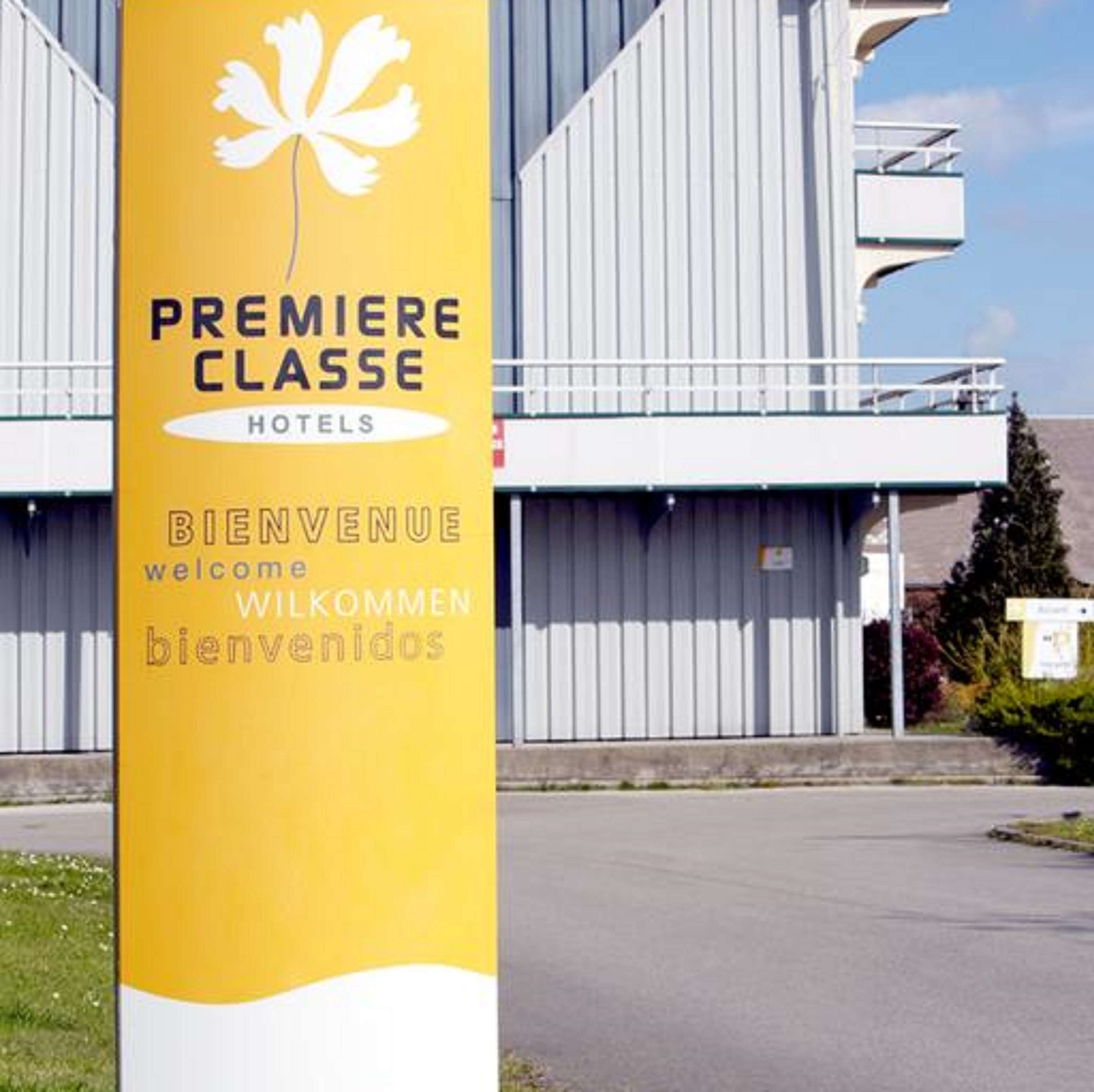 Una señal amarilla con un logo de hoja blanca y el texto 'PREMIERE CLASSE HOTELS' y 'BIENVENUE welcome WILKOMMEN bienvenidos'.
