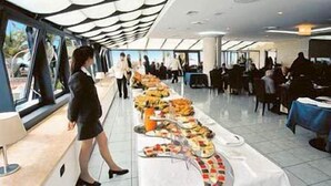 A modern indoor space with a long table displaying various food items, a woman walking, and a crowd in the background. - Hotel Gli Dei (Pozzuoli)