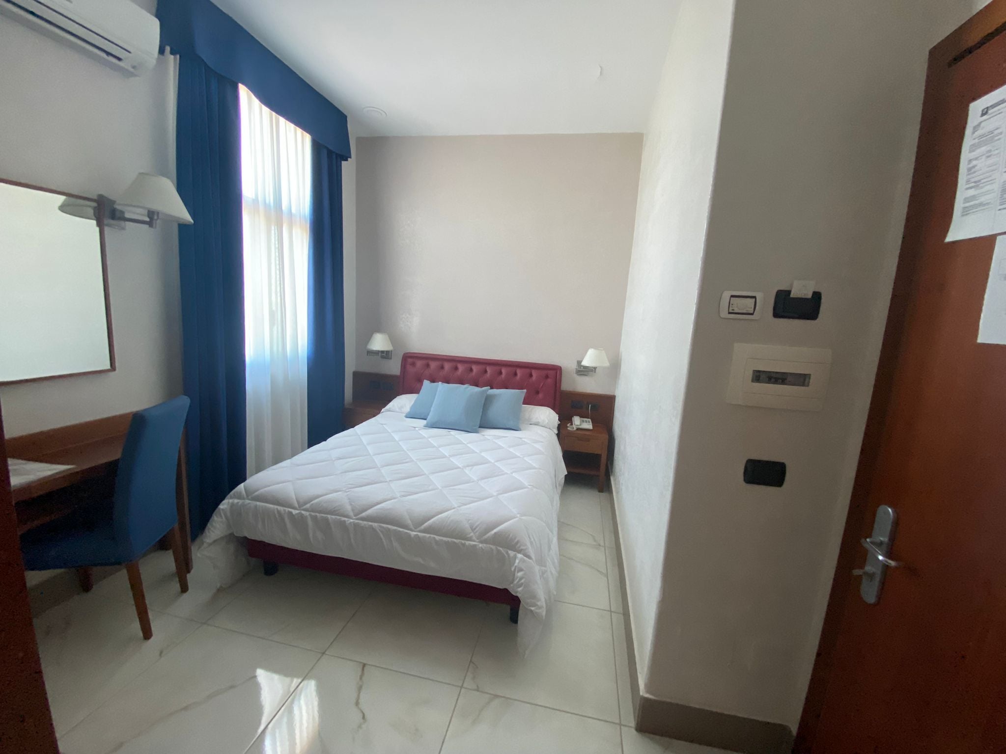 A hotel room with a bed, a desk, a chair, a window with curtains, and a door.