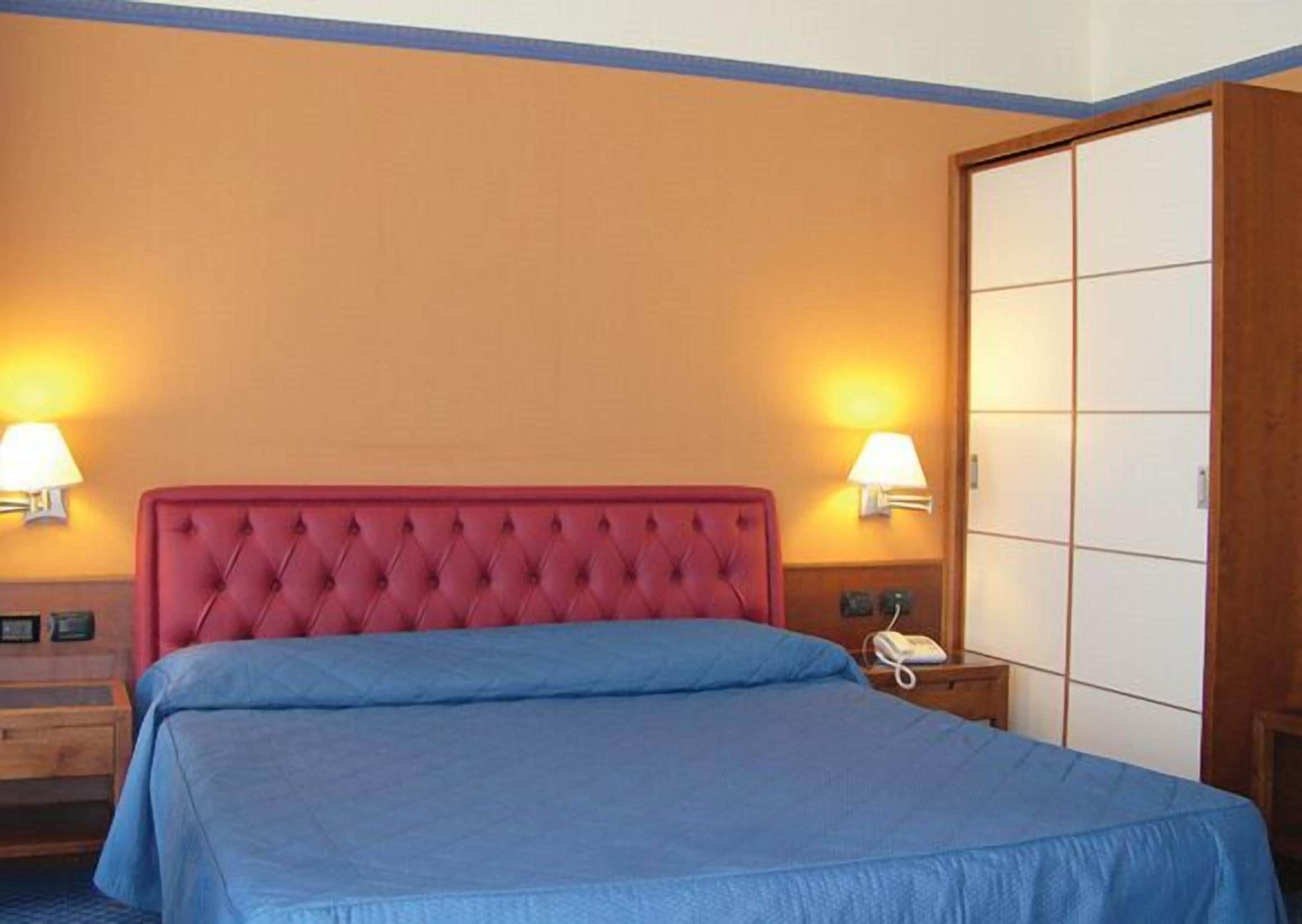 Een hotelkamer met een rode gewatteerde hoofdbord, een blauw beddegoed, twee nachtlampjes, een houten nachtkastje en een houten garderobe.