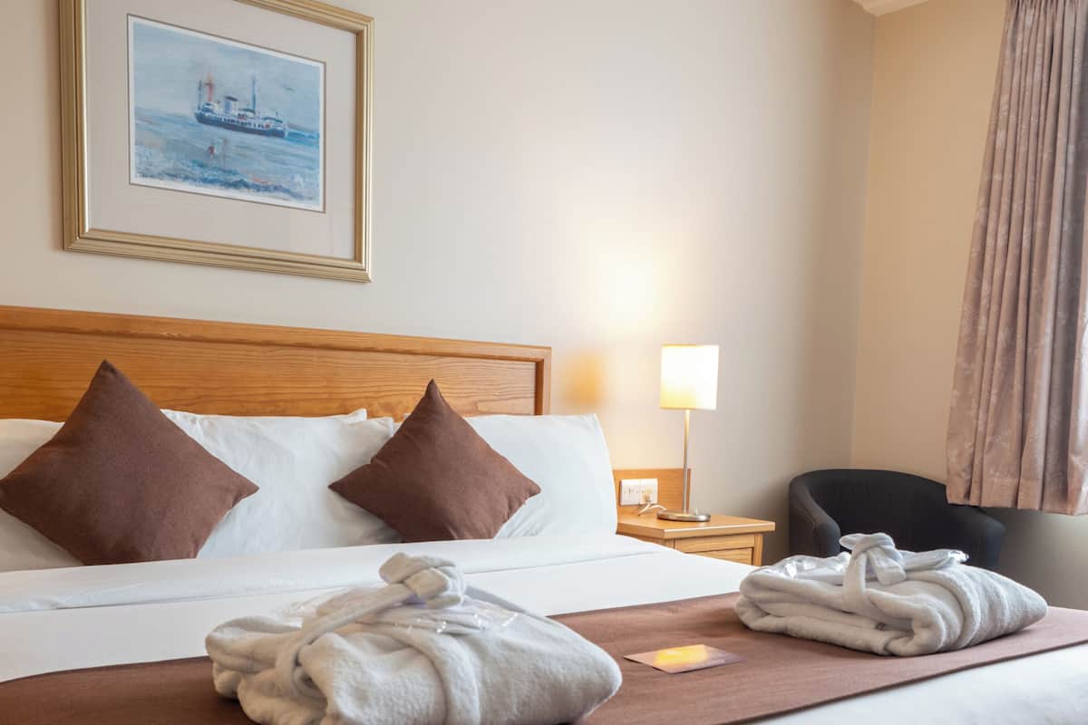 Habitació d'hotel amb un llit gran, dos coixins, un capçal de llit, una taula de nit amb una làmpada, una cadira, un quadre a la paret i una finestra amb cortines.