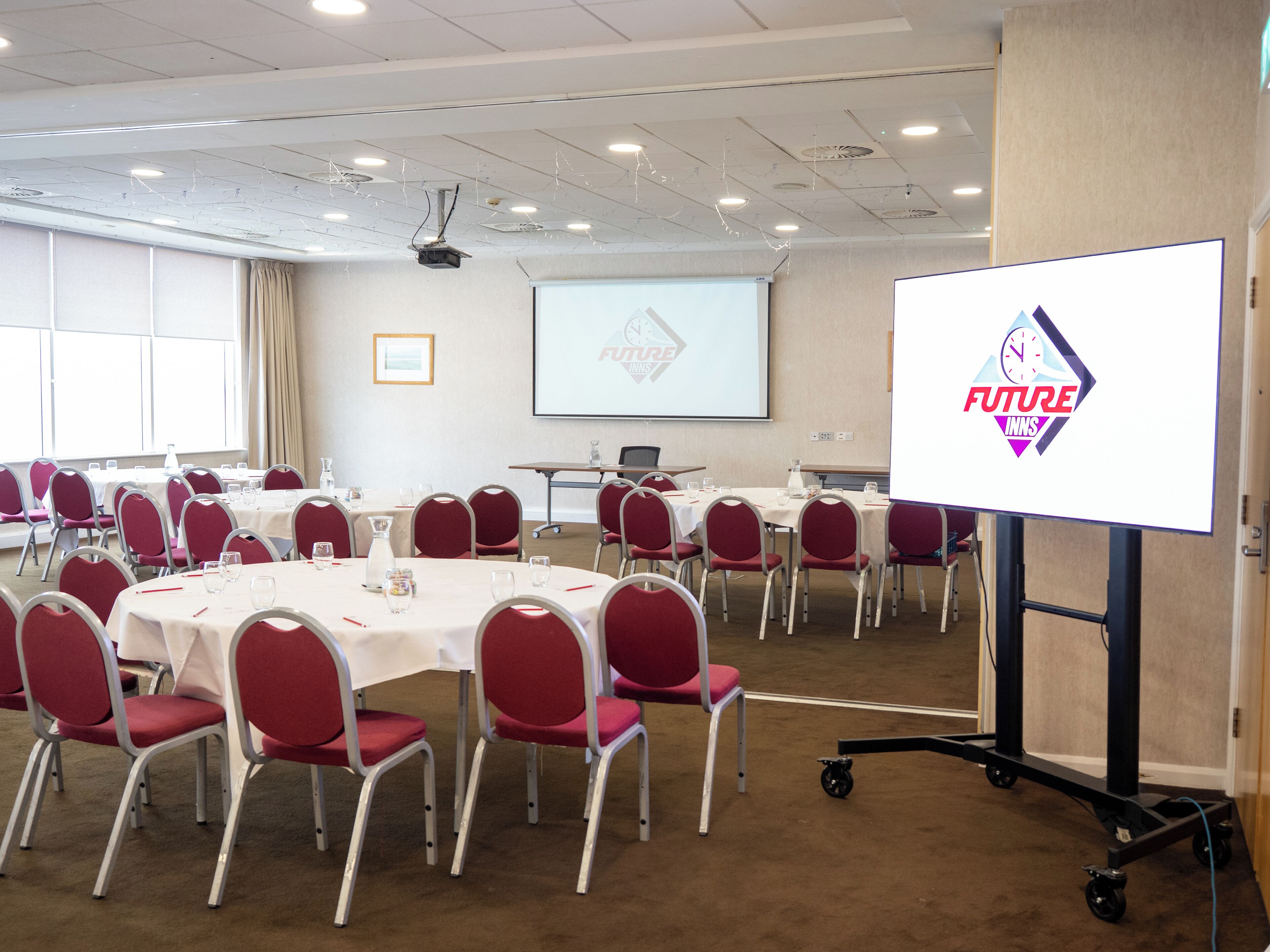 Une salle de conférence équipée de tables rondes, de chaises rouges et d’un écran de projection affichant un logo.