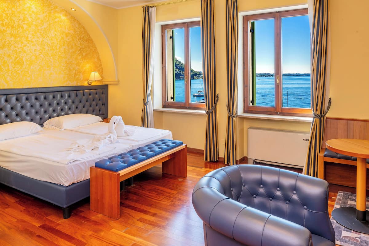 A hotel room with a bed, a bench, a chair, a table, and a view of the sea.