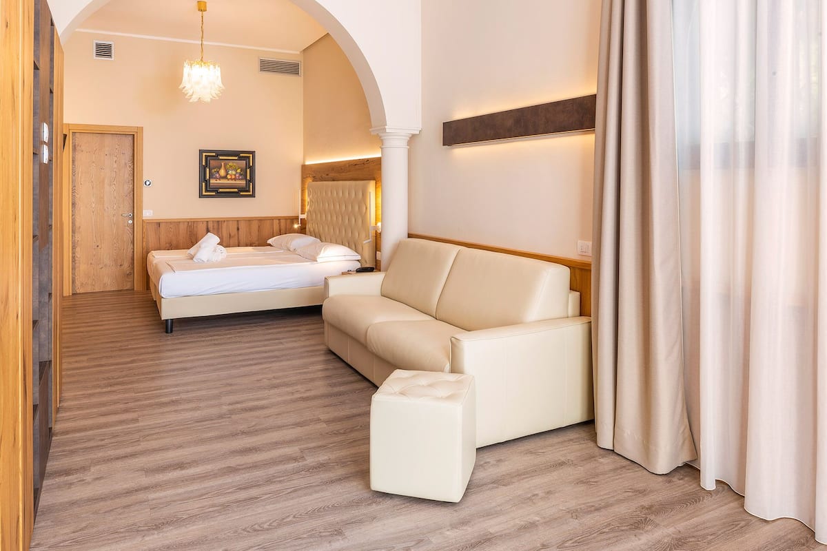 A hotel room with a bed, a sofa, and a small ottoman. There is a wooden door and a framed picture on the wall.