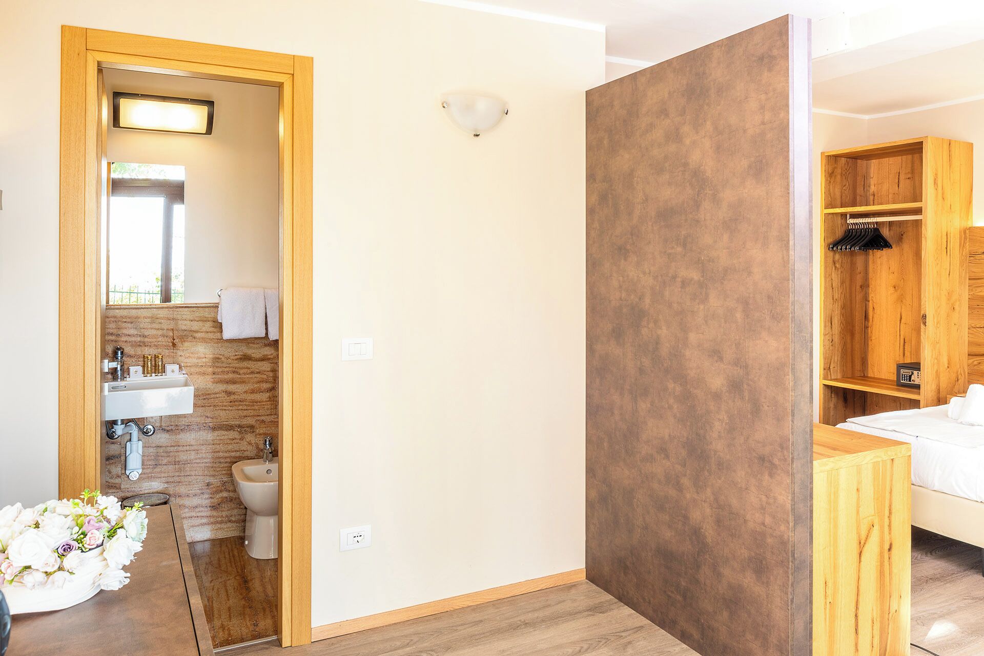Ein modernes Hotelzimmer mit Holzboden, einer Holzwand, einem Badezimmer mit Waschbecken und Bidet sowie einem Kleiderschrank mit Kleidungsstücken.