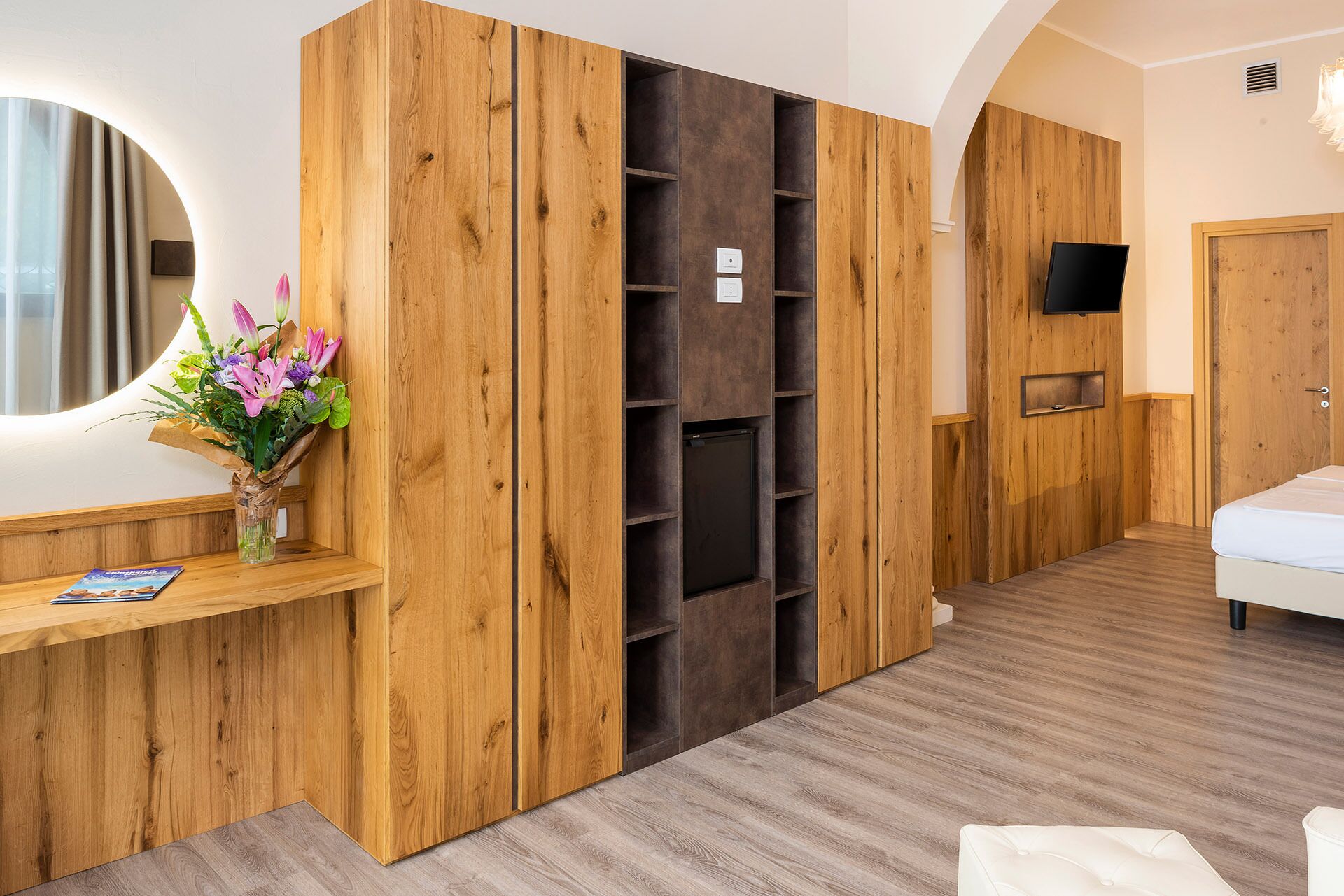 Una habitación de hotel con un armario de madera, un escritorio pequeño con un florero y una cama, además de un televisor montado en la pared.