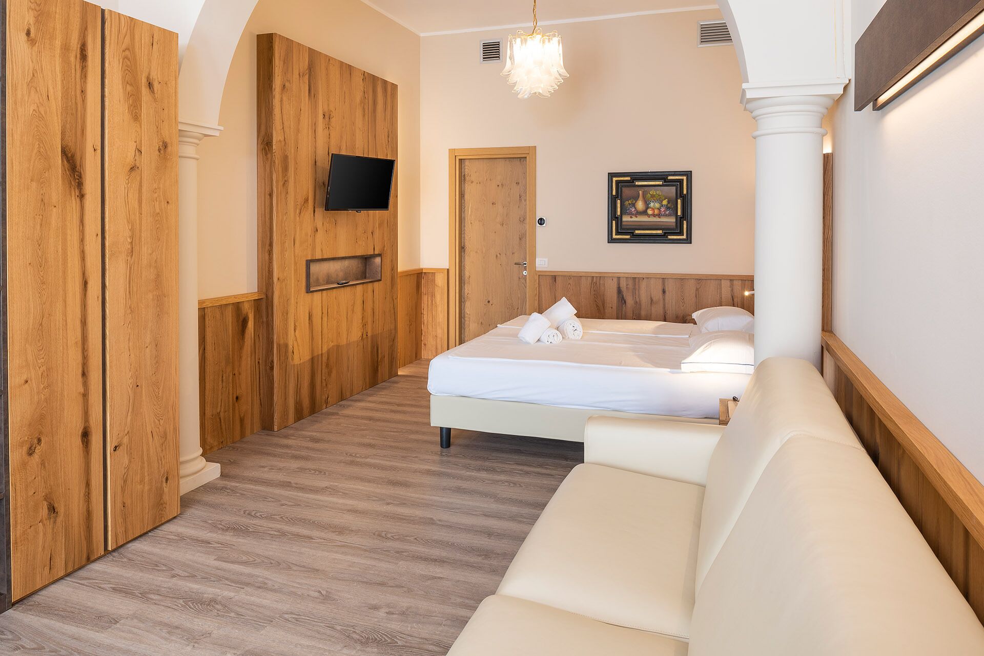 Une chambre d’hôtel avec un mur lambrissé en bois, un lit avec du linge de maison blanc, une table de chevet, un luminaire suspendu et un tableau encadré au mur.