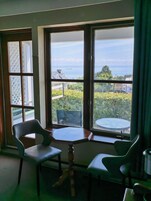 Um quarto com uma mesa, duas cadeiras, uma janela com vista para o mar e edifícios, e uma porta com painel de vidro trabalhado.