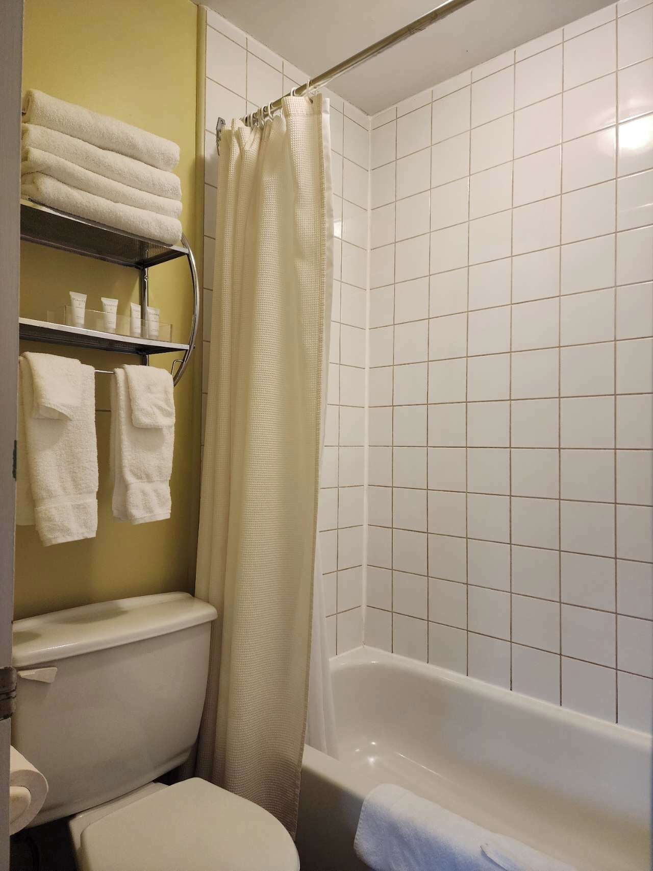 Un baño con inodoro, bañera y cortina de ducha. Hay toallas y artículos de aseo personal en un estante.