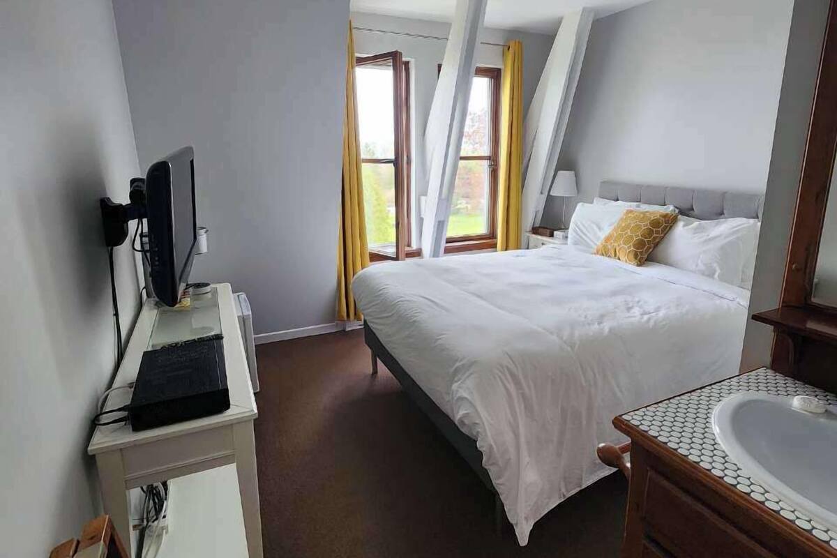 Habitación de hotel con cama, escritorio con computadora, baño con lavamanos y ventanas grandes con cortinas.