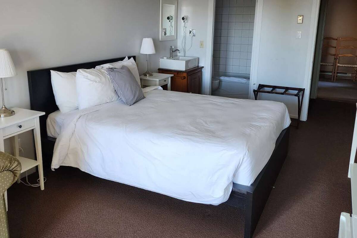 Une chambre d’hôtel avec un grand lit, une table de chevet avec une lampe, une petite table avec un miroir, une salle de bain visible à travers une porte ouverte et une chaise dans un coin.