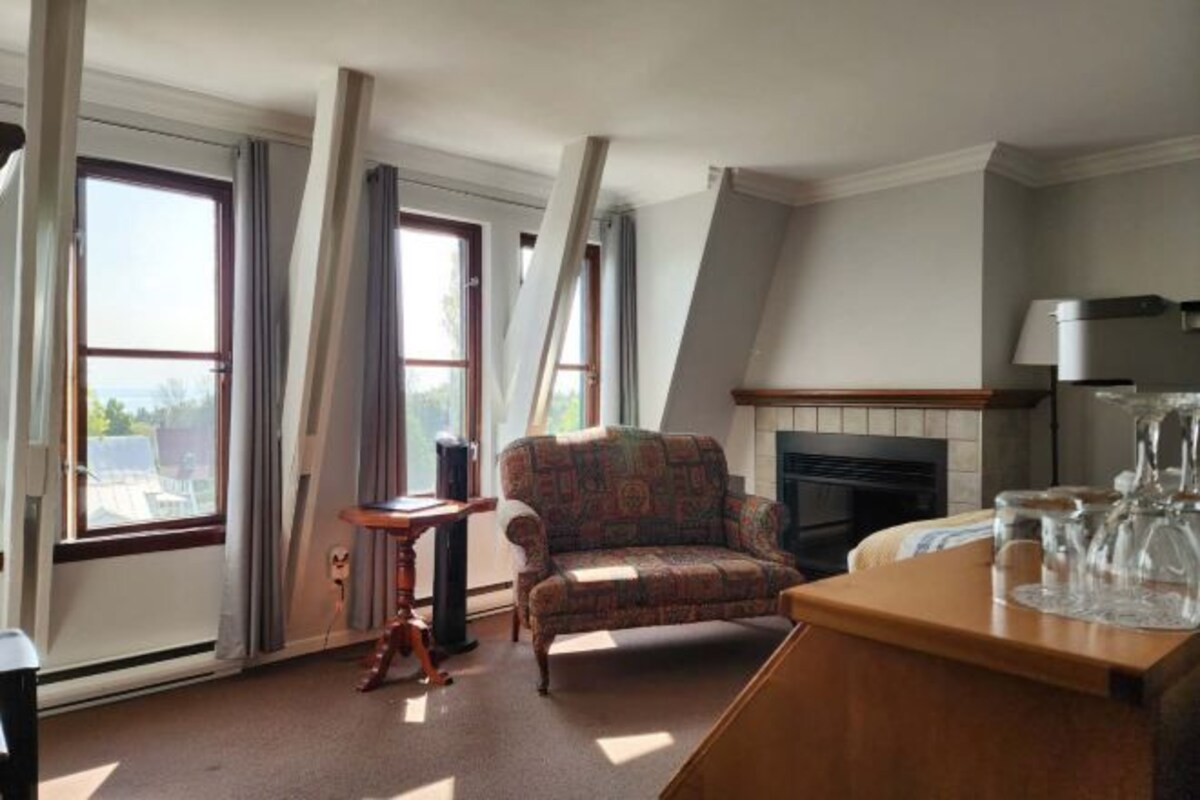 A living room with a fireplace, a patterned armchair, a small table, and a view through large windows.