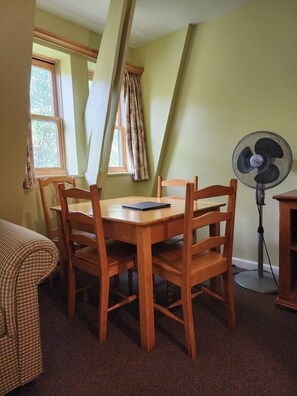 Un comedor con una mesa y sillas de madera, un ventilador de pie y una ventana con cortinas.