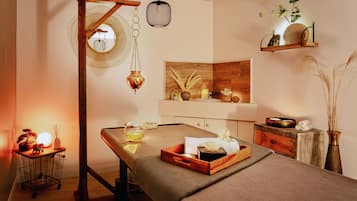 Una habitació d'spa amb un llit de massatge, una taula auxiliar de fusta amb una làmpada, una estant amb objectes decoratius i un mirall a la paret.