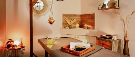 A spa room with a massage table, a wooden side table with a lamp, a shelf with decorative items, and a mirror on the wall.