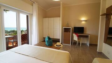 A hotel room with a bed, a desk, a TV, and a view of the sea.