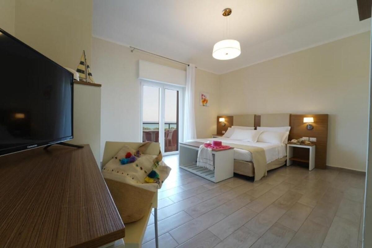 A hotel room with a bed, bedside tables, a TV, a sofa, and a balcony.
