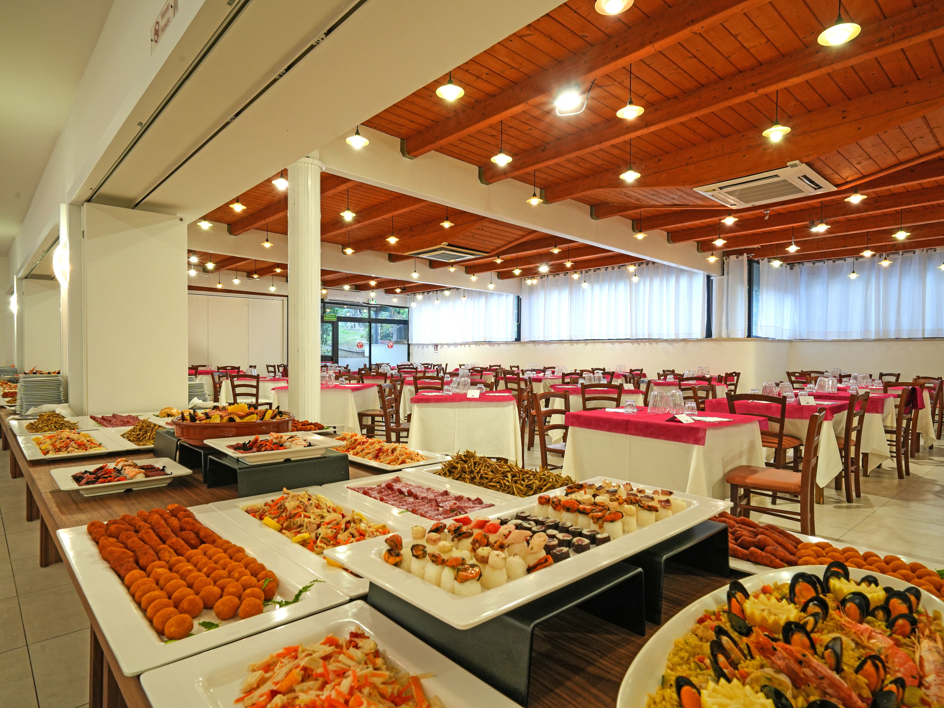 Un menjador tipus bufet amb diversos plats i taules, organitzat en una sala espaiosa amb bigues de fusta i grans finestres.