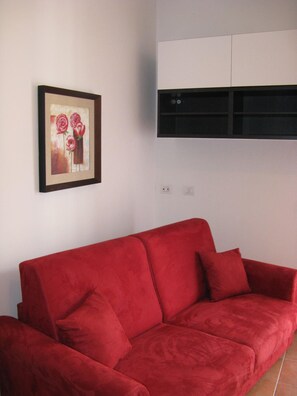 Um sofá vermelho em um quarto com parede branca e um quadro com flores.