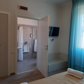 Quarto de hotel com cama, chuveiro, sanita e televisão fixada na parede, além de janela com cortinas.