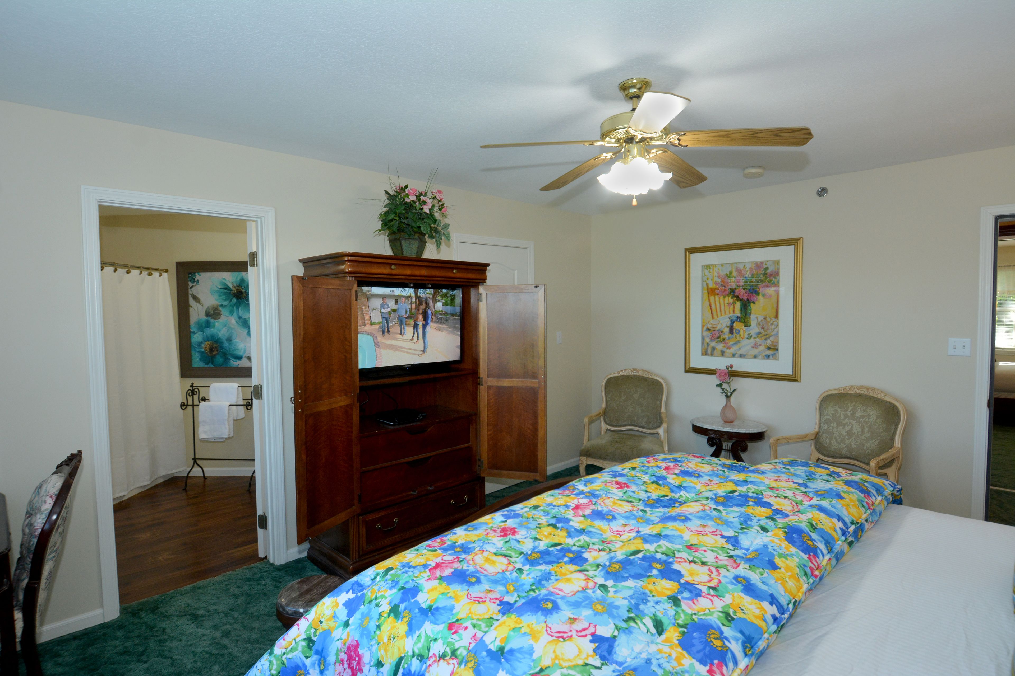 A bedroom with a bed, a ceiling fan, a dresser, a television, two chairs, and a painting on the wall.