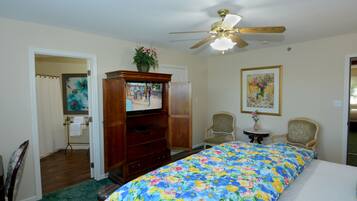 A bedroom with a bed, a ceiling fan, a dresser, a television, two chairs, and a painting on the wall.