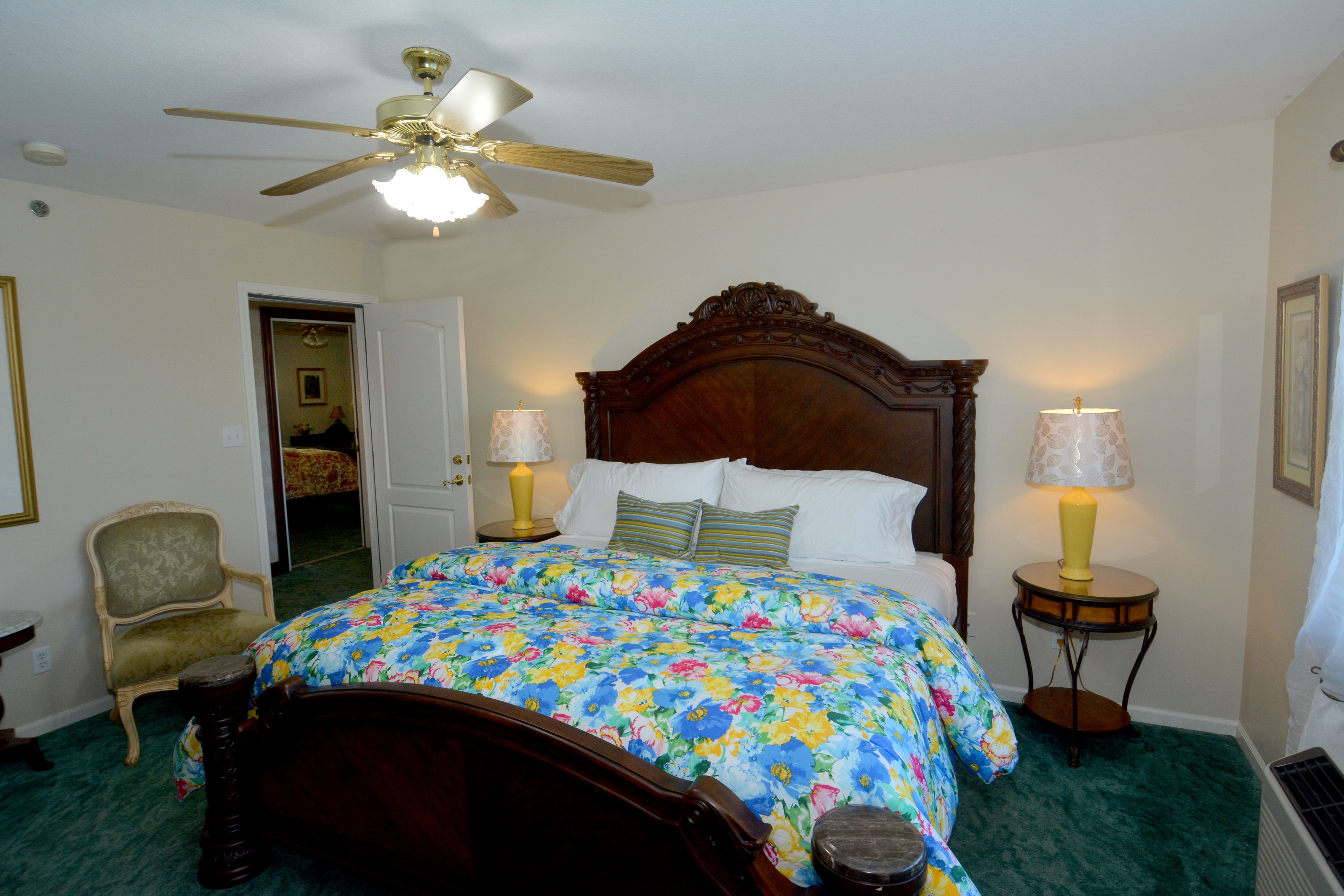 A bedroom with a wooden bed, floral bedspread, two bedside lamps, a chair, and a ceiling fan.