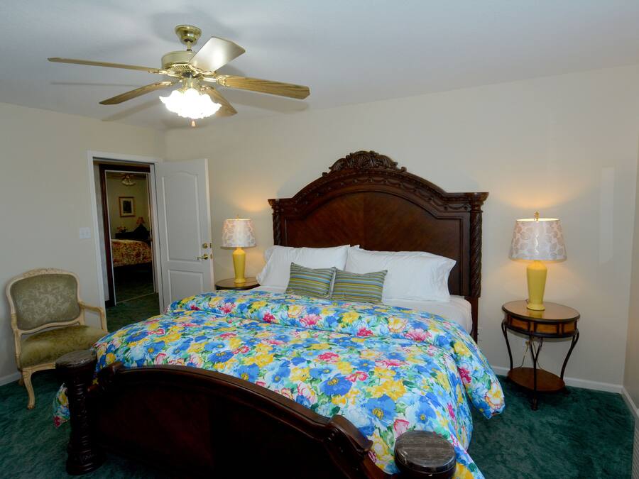 A bedroom with a wooden bed, floral bedspread, two bedside lamps, a chair, and a ceiling fan.