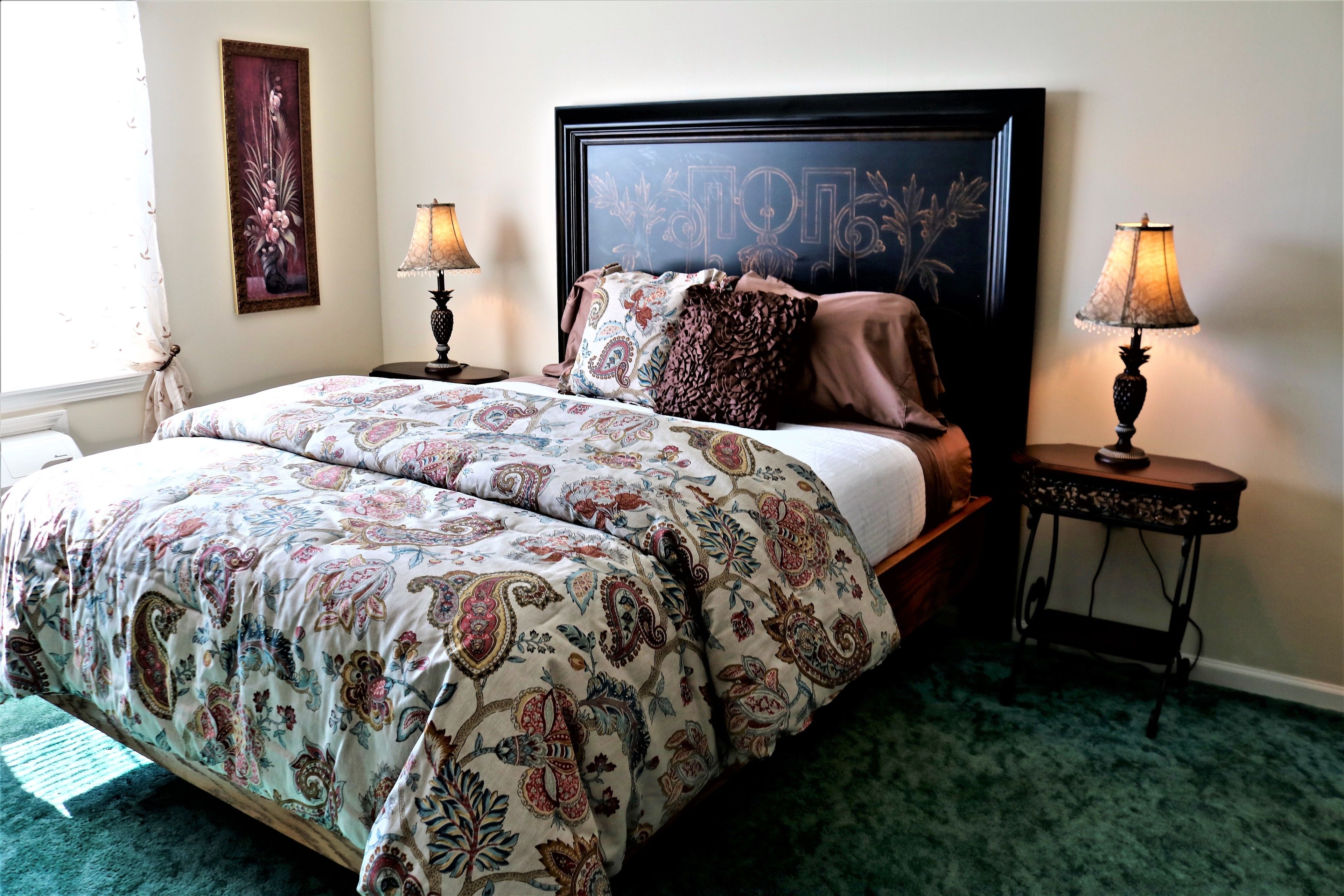 A bedroom with a large bed, two bedside tables with lamps, a framed artwork on the wall, and a patterned bedspread.