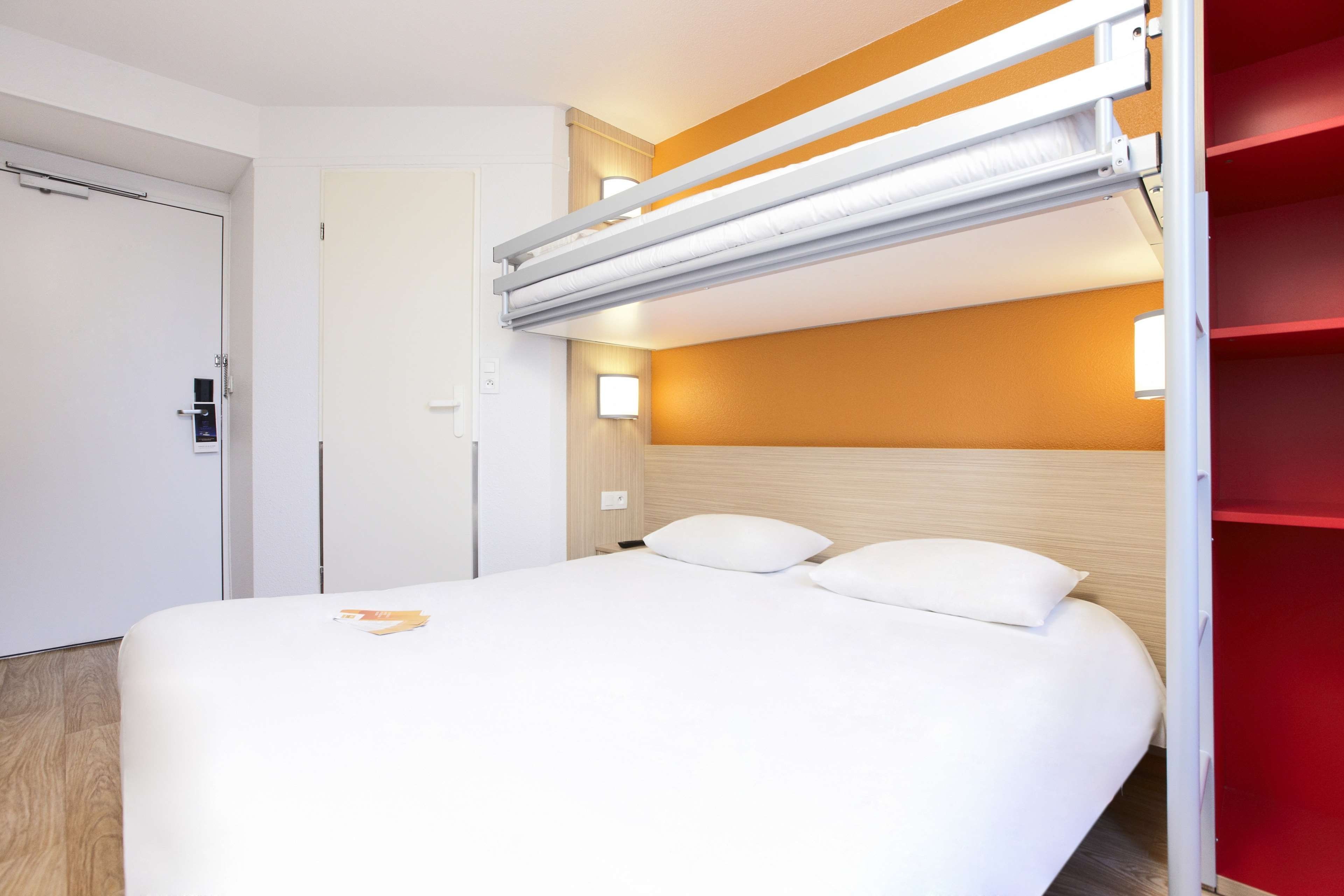 A hotel room with a bunk bed, a white bed, and a red shelving unit.