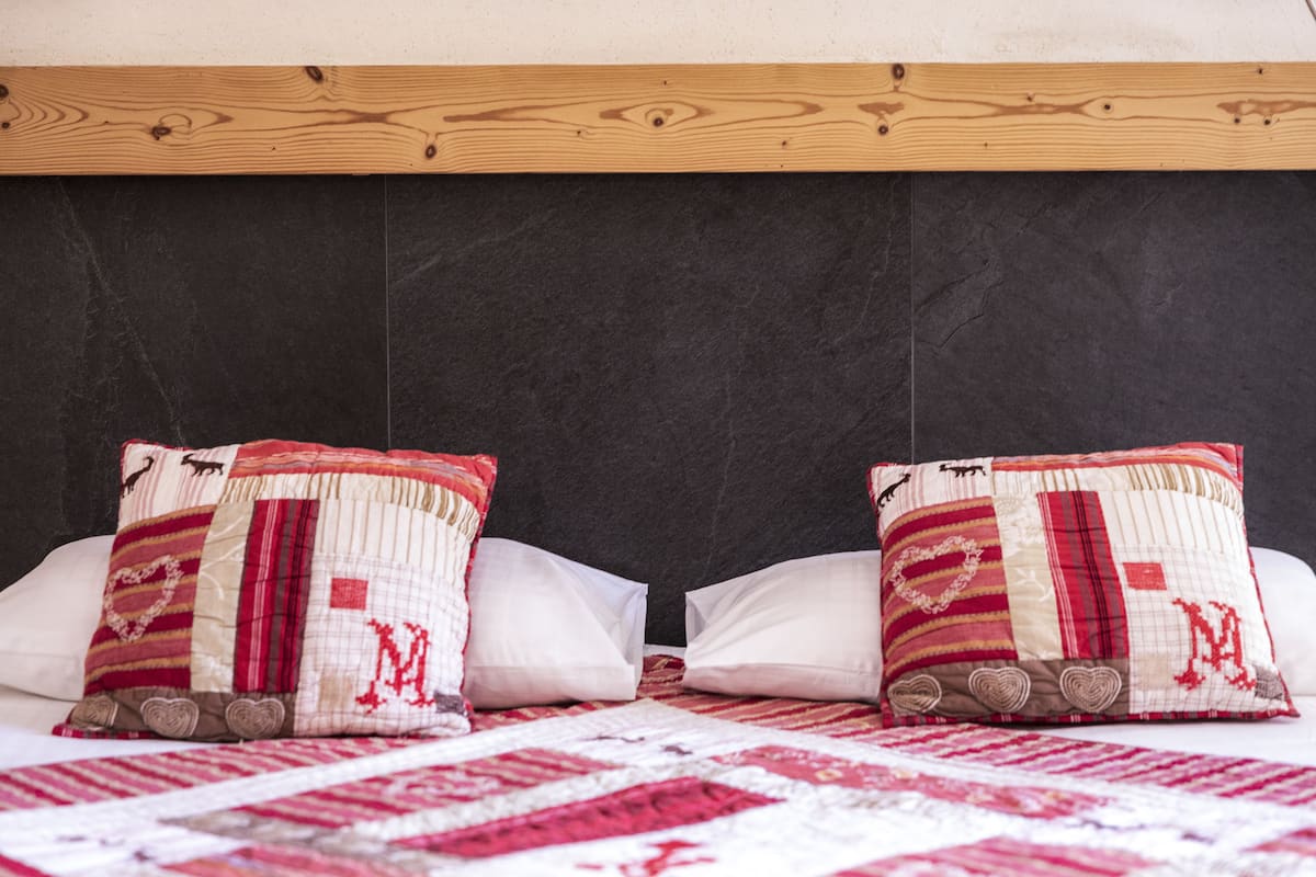 Una cama con cabecera oscura, dos almohadas con estampado rojo y beige, y una colcha roja y blanca.