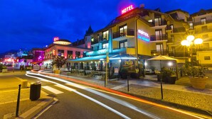 Ein Hotel mit einem auffälligen 'HOTEL'-Schild, ein Pizza-Restaurant namens 'PIZZA ERIA GLASER' gegenüber und eine gut beleuchtete Straße mit Lichtspuren vorbeifahrender Fahrzeuge.