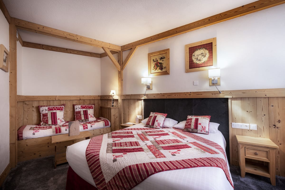 Un dormitorio con una cama grande, dos camas más pequeñas, vigas de madera, cabecera, luces fijadas a la pared y cuadros enmarcados.