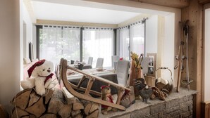 Ein gemütliches Zimmer mit Holzbalkendecke, einem großen Fenster, einem Esstisch mit Stühlen und einem Kamin mit Holzscheiten und einem Teddybären.