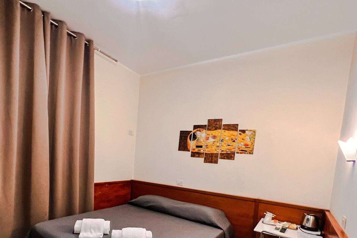 A hotel room with a bed, a small table with a kettle, and a wall-mounted artwork.