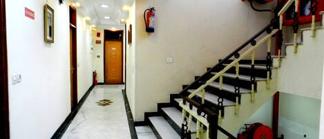 A hotel hallway with a marble floor, a staircase with black and white railings, and a fire extinguisher mounted on the wall.