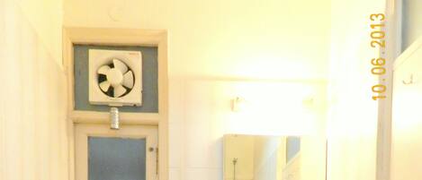 A small bathroom with a toilet, sink, and mirror. There is a window above the toilet and a small shelf with a fan.
