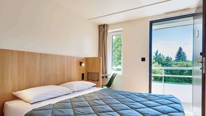 Una habitación de hotel moderna con una cama grande, cabecera de madera, un escritorio pequeño y un ventanal corredero que da a un balcón con vistas a árboles.