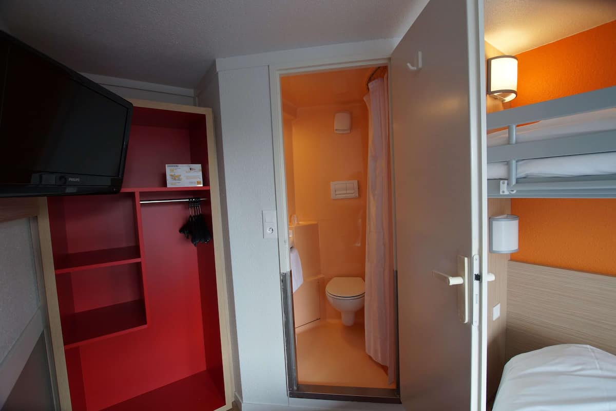 A compact hotel room with a red storage unit, a single bed, a shower area, and a television mounted on the wall.