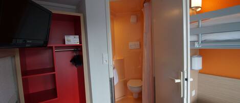 Habitación de hotel compacta con un mueble de almacenamiento rojo, una cama individual, zona de ducha y televisor montado en la pared.