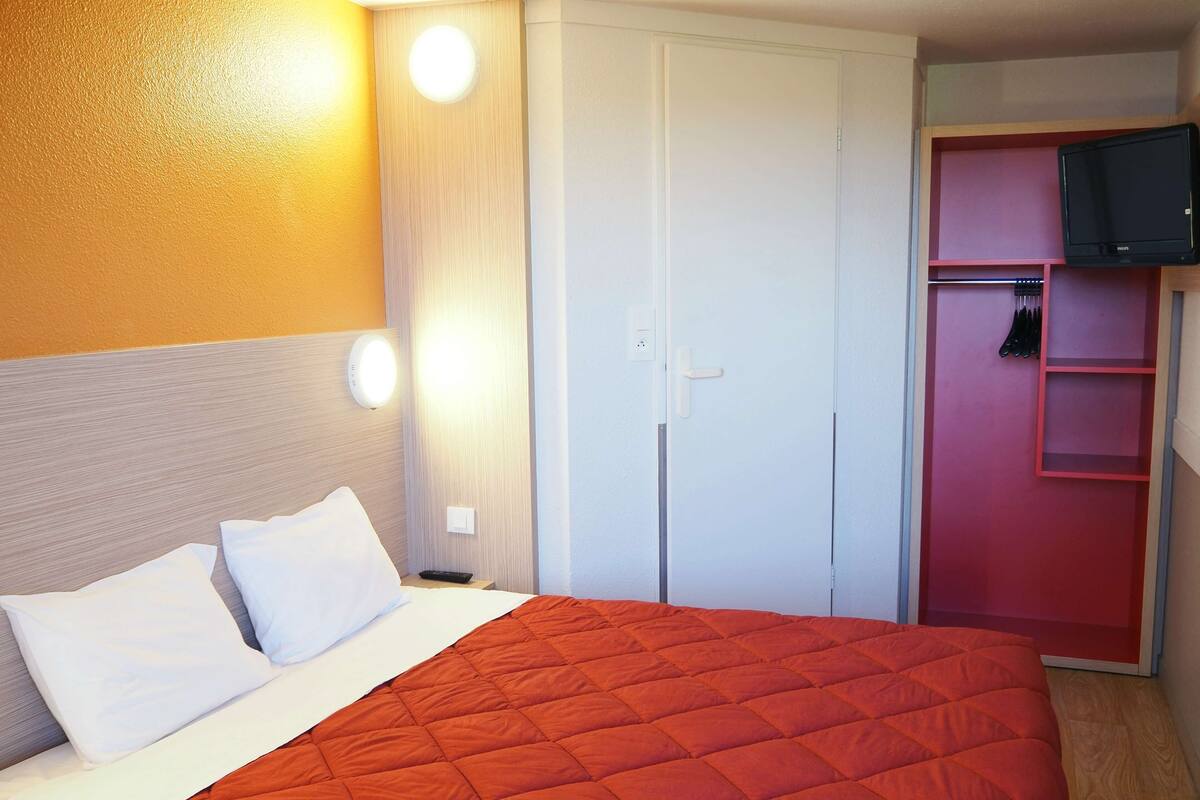 A hotel room with a bed, a wardrobe, a television, and a wall-mounted light fixture.