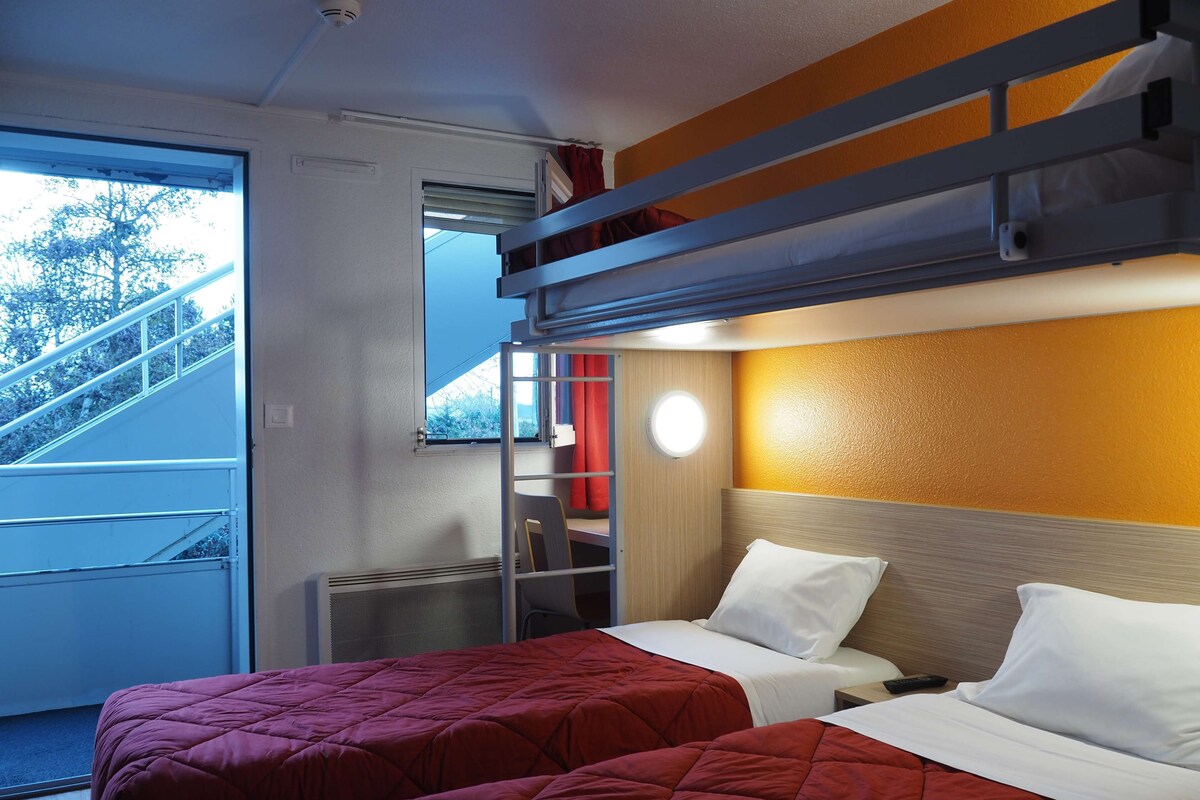 Habitación de hotel con dos camas, una litera, una ventana con vistas a una escalera y una lámpara de pared redonda.