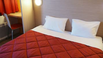 Une chambre d’hôtel avec un lit, deux oreillers, une couvre-lit rouge matelassée, un bureau avec une chaise et une lampe fixée au mur.